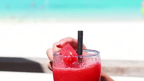 a hand stirs a vibrant watermelon drink with a straw, set against a bright beach backdrop.