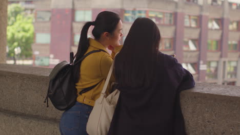 Rear-View-Of-Two-Young-Female-Friends-Visiting-The-Barbican-Centre-In-City-Of-London-Looking-At-Mobile-Phone