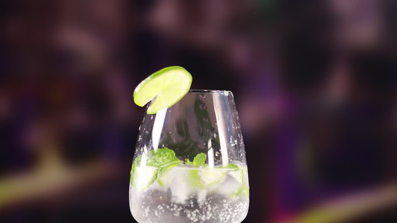 A mojito cocktail with lime and mint in a clear glass against a blurred, colorful background