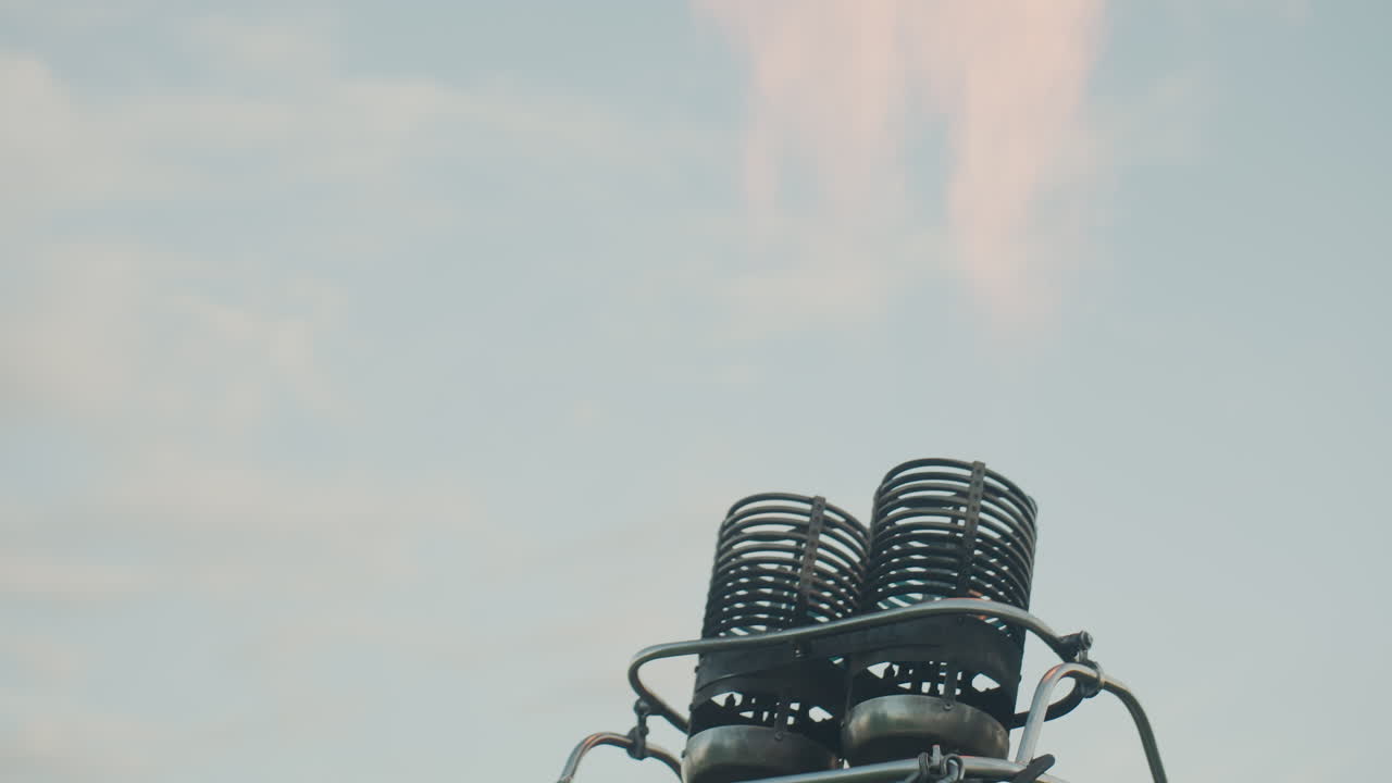 close up of metal burner coils igniting twin orange flames above wicker basket of hot air balloon causing fire to flicker through spirals against pale sky at dusk during flight launch preparation