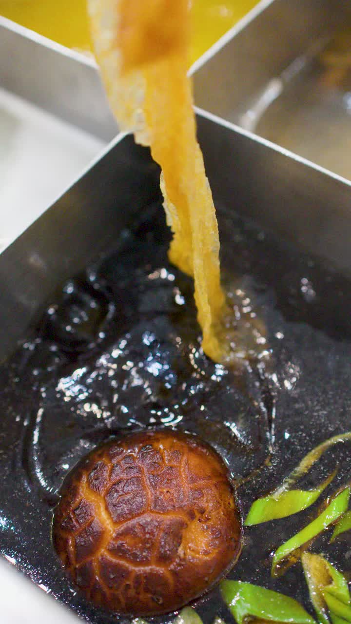 Close-up shot of chopsticks picking up tofu skin from dark broth in a divided pot