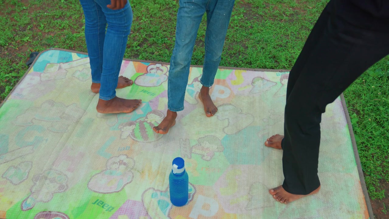 Mère noire pieds nus enseignant à son enfant sur un tapis coloré dans le jardin, jean en denim, bouteille d'eau à proximité, instruction ludique, après-midi chaleureuse, moment authentique de lien familial et de guidance