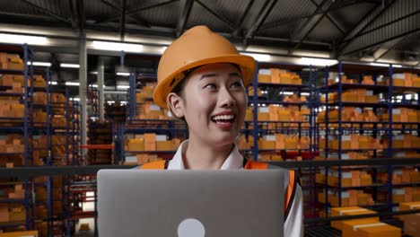 close up de ingeniera asiática con casco de seguridad mirando una computadora portátil y mirando a su alrededor mientras está de pie en el almacén con estantes llenos de bienes de entrega