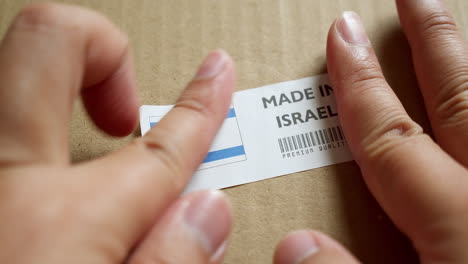 Manos-Aplicando-La-Etiqueta-De-La-Bandera-Hecha-En-Israel-En-Una-Caja-De-Envío-Con-Un-Código-De-Barras-De-Primera-Calidad-Del-Producto