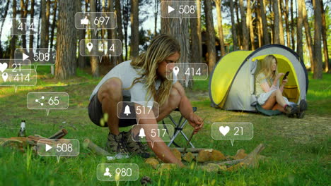 male crouching arranging campfire wood in forest clearing with floating social media ads