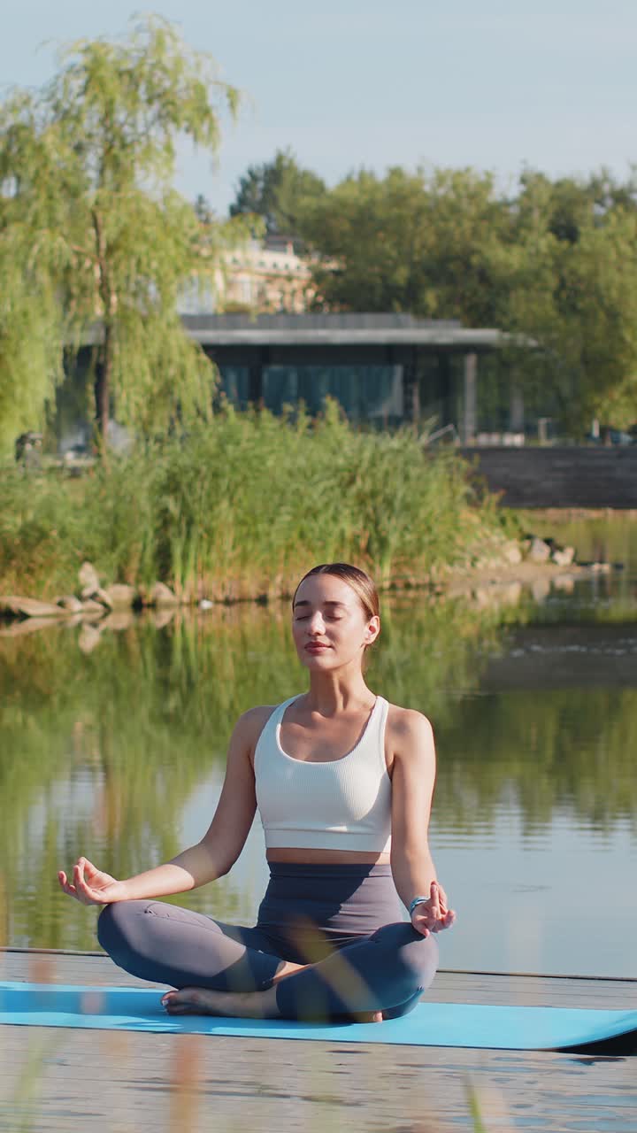 Caucasian young woman sitting on mat in lotus position relaxing near pond in park on summer day