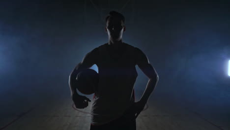 the basketball player stands on a dark playground and holds the ball in his hands and looks into the camera in the dark with a backlit in slow motion and around smoke