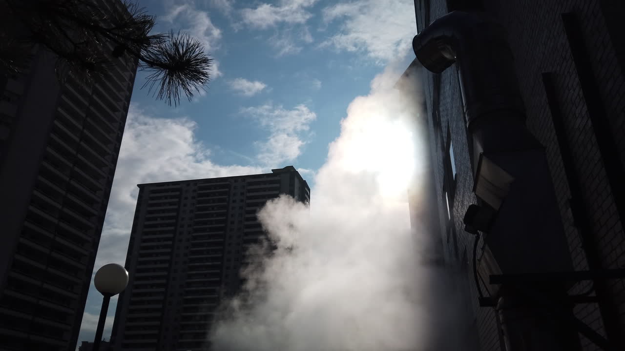 Steam escaping a tall metal vent outside towering high rise buildings, low angle wide shot