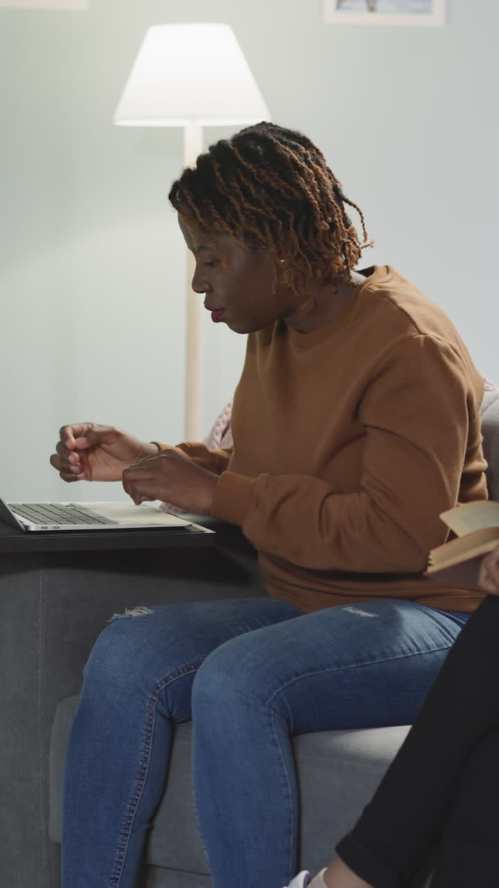 African American and Indian women prepare for student work sitting on sofa in hostel room. Multiracial girl friends read books and use laptop for studying