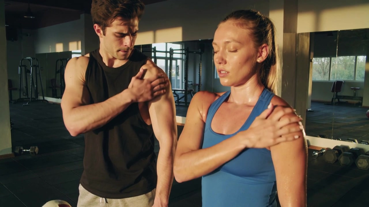 Uomo con canotta nera che massaggia la spalla destra di una donna dopo l'allenamento in palestra, con manubri.