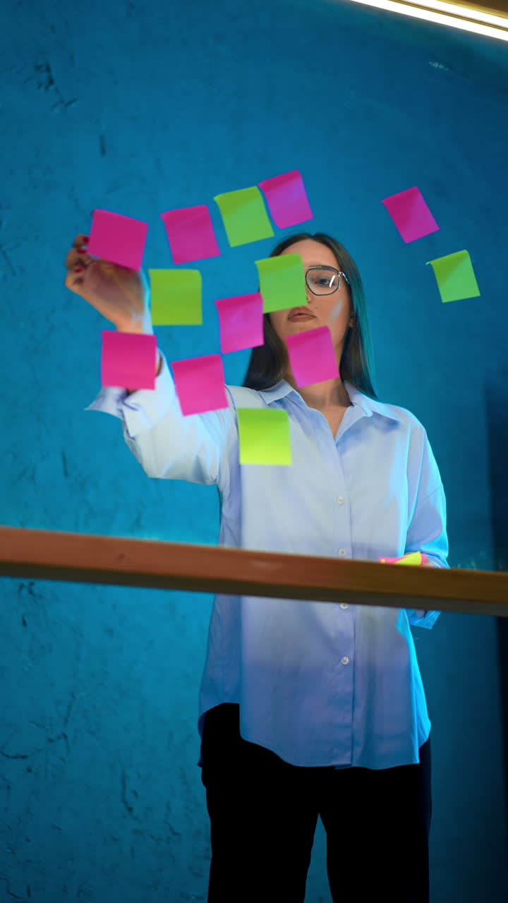 Lady in white shirt and glasses put sticky notes on the glass board. Businesswoman arranging plans, memos on the glass wall. Vertical video.