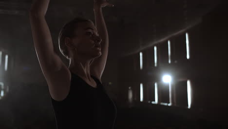 Slow-motion:-Diligent-young-graceful-ballerina-dancing-elements-of-classical-ballet-in-the-dark-with-light-and-smoke-on-the-background.-Beautiful-young-ballerina-in-darkness.