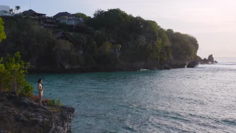 aerial view of young girl clad in bikini standing on clifftop at beachside and enjoying beautiful landscape