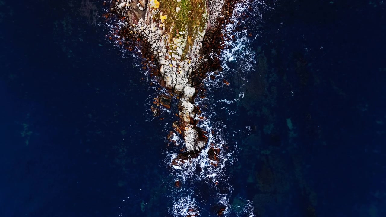 Drone view of coastal rock formation. Small grassy point with a hut sits above jagged stones where waves break along the shore.