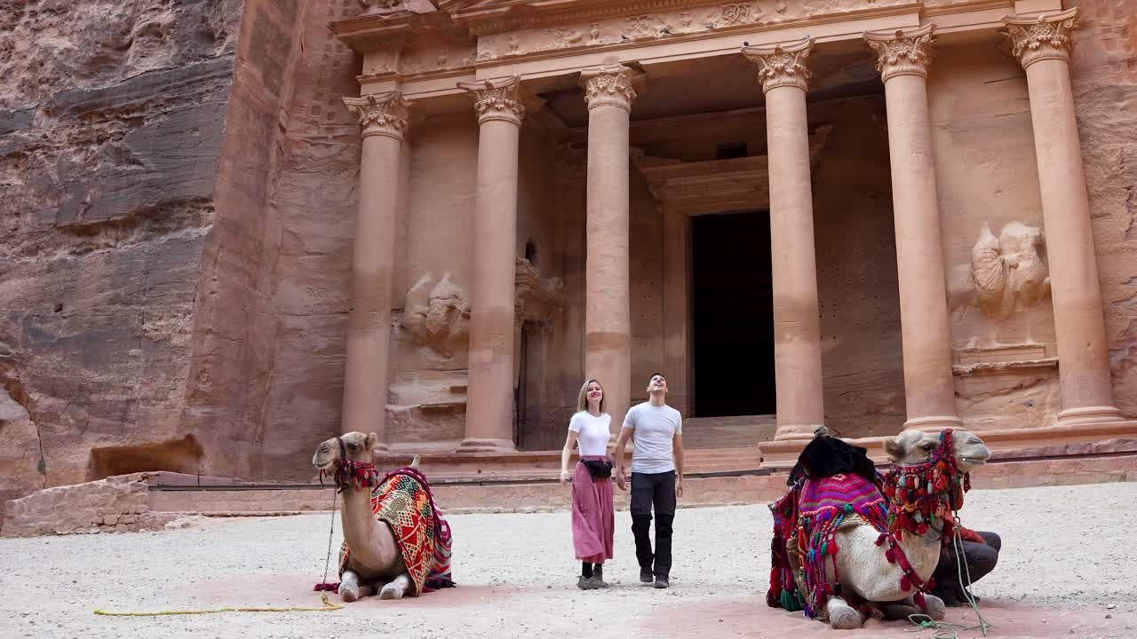 Young couple walks hand in hand toward the ancient Treasury monument in Petra, Jordan, with camels resting in front of the carved façade, capturing travel, culture, and Middle Eastern heritage