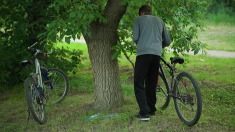 vista trasera de un niño que lleva una camiseta gris estacionando su bicicleta cerca de un árbol grande mientras mueve las ramas, una segunda bicicleta se encuentra cerca en una zona de hierba, rodeada de exuberante vegetación