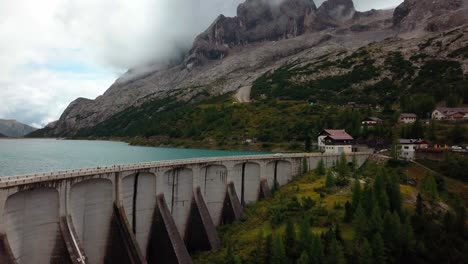 Wall-of-Lake-Fedaia-Dam-in-the-Dolomite-mountain-area-of-northern-Italy-with-people-and-cars-moving-on-the-top,-Aerial-Drone-dolly-in-reveal-shot