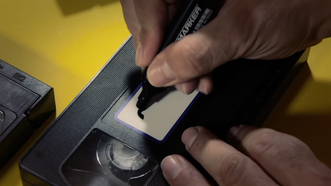 Writing the text 2000's on the white label of an old VHS videotape , with a marker