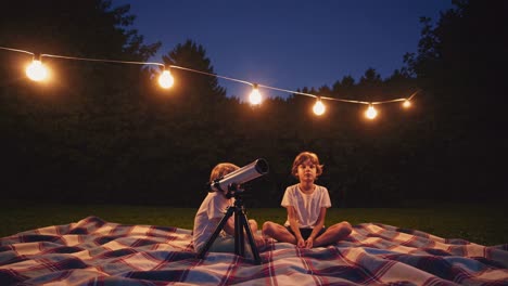 two children stargazing with a telescope on a plaid blanket under string lights