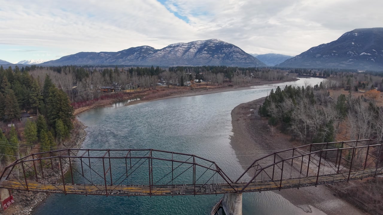Aerial view of a broad river snaking through a forested valley, with a rustic steel bridge in the foreground and snow-dusted mountains rising under a pale sky.