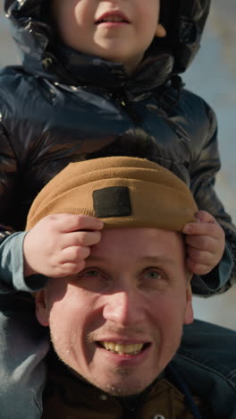 un padre lleva a su hijo pequeño en los hombros mientras señala algo interesante, cubriendo los ojos de su padre con las manos y ajustando la gorra
