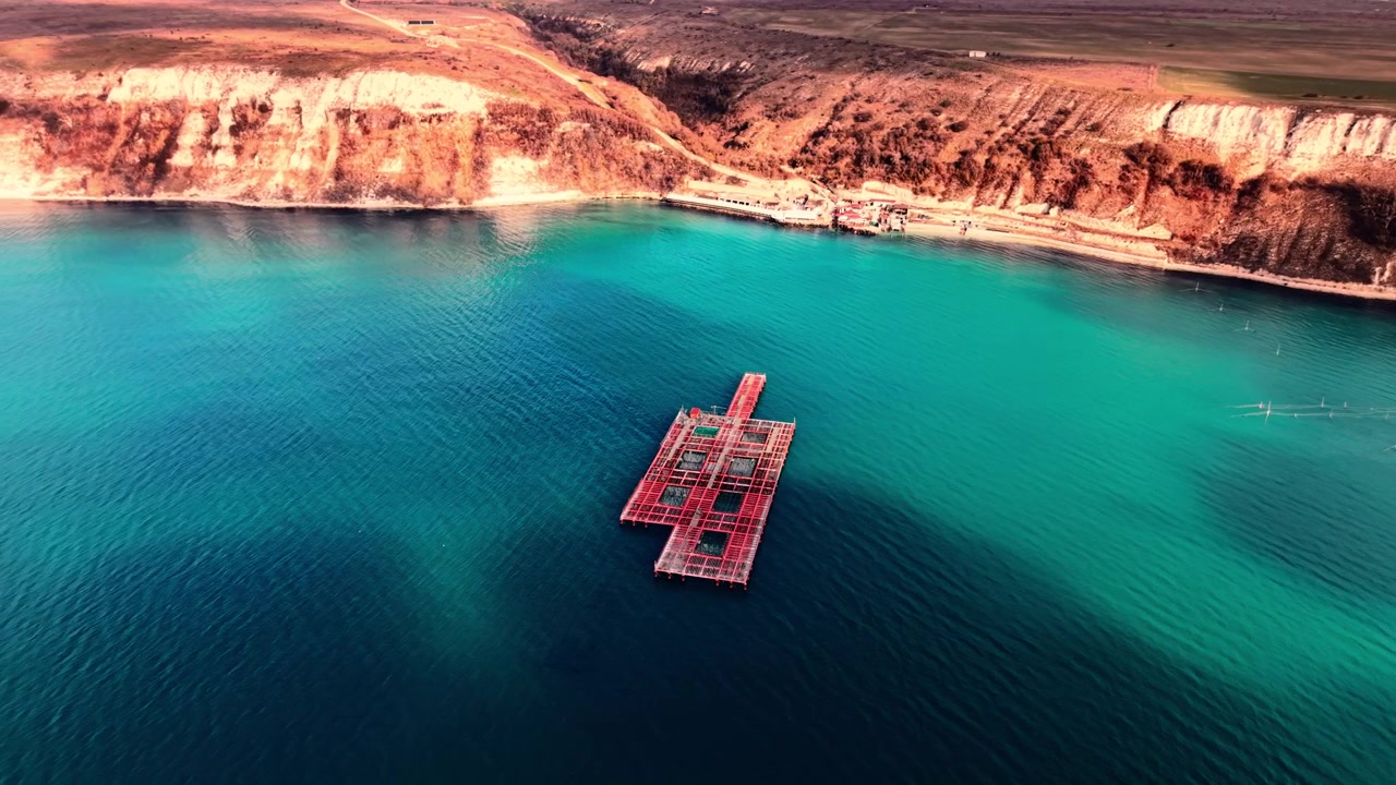 Fish farm platform in coastal bay with white chalk cliffs. Aerial view of red aquaculture structure on vivid turquoise water enclosed by eroded limestone headland and farmland above.