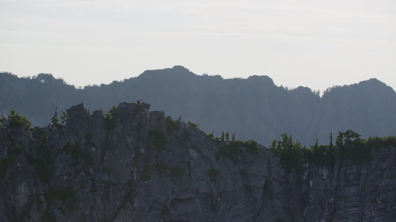 모험 사진작가는 풍경을 볼 수 있는 거대한 산 능선에 홀로 서 있고, 팀 더칸 극단적인 넓은 추적 헬리콥터는 시계 반대 방향으로 느린 궤도를 돌고 있습니다.