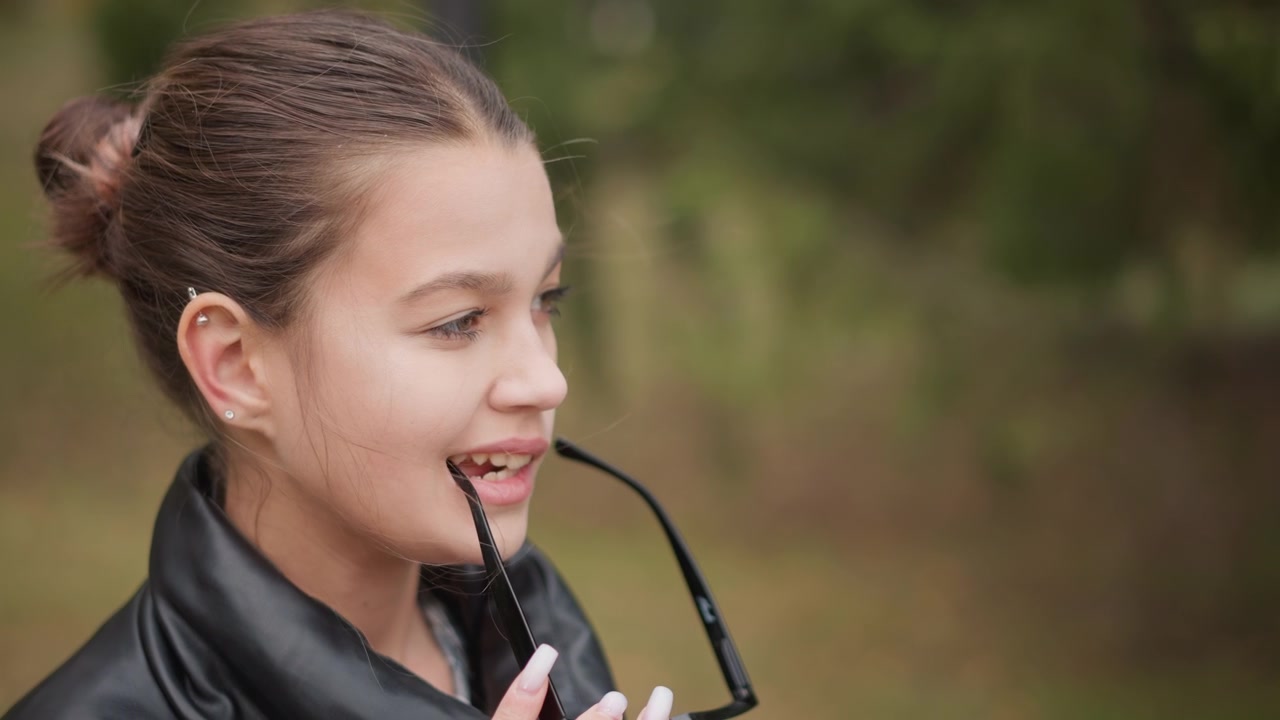 Primo piano di una giovane donna che tiene occhiali neri in un ambiente esterno con vegetazione. La donna indossa una giacca di pelle e ha piercing all'orecchio visibili, mantenendo la stessa posa contro uno sfondo naturale sfocato