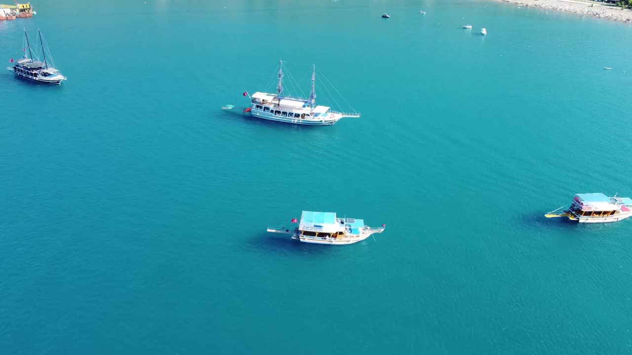 Aerial drone view showing turquoise water with multiple boats near coastal hills in Antalya, capturing calm sea surface, clear summer light, peaceful shoreline, and scenic blue horizon