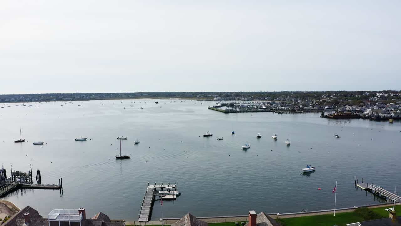 Dron volando a la izquierda sobre el puerto de Nantucket en Massachusetts, revelando veleros dispersos, muelles en funcionamiento y la tranquila ciudad costera extendiéndose a lo largo de la costa en una amplia toma aérea marítima.