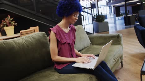 mujer de negocios biracial con afro azul y usando computadora portátil en la oficina, cámara lenta