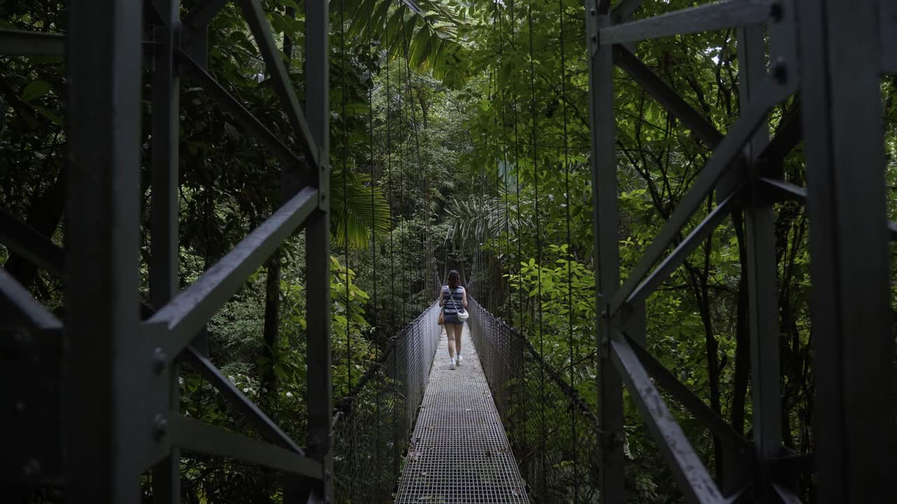 Handheld shot of the Mistico hanging bridge in La Fortuna, Costa Rica. A young woman walks away from the camera on the long gray suspension bridge, surrounded by lush green foliage on a summer morning