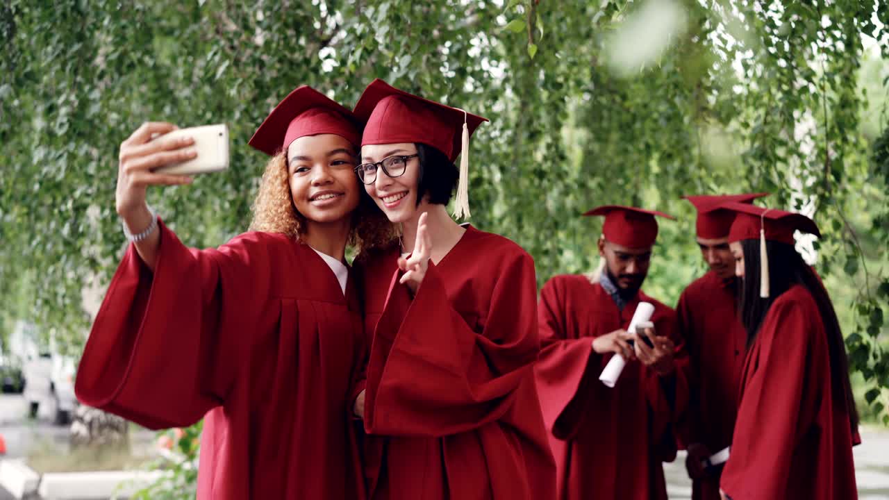 Pretty female graduates are taking selfie with diploma scroll using smartphone, young women are posing with other students moving and talking in background.