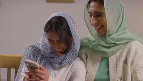 Muslimische-Mutter-Und-Tochter-Lachen-über-Ihr-Mobiltelefon-Und-Sitzen-Zu-Hause-Am-Muslimischen-Familientisch-1