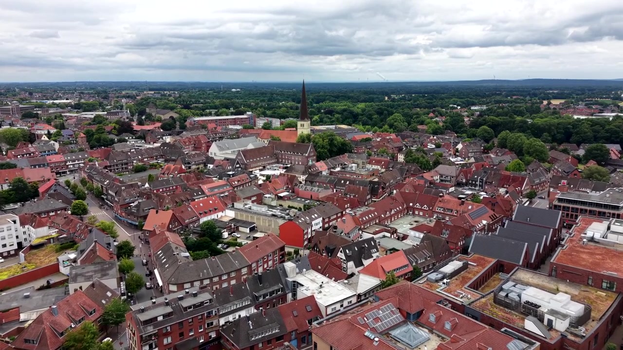 Campanario de iglesia histórico se eleva sobre tejados de ladrillo rojo en el casco antiguo de Dülmen, Renania del Norte-Westfalia, Alemania, rodeado de bosque verde y campo más allá del centro de la ciudad. Toma aérea amplia de dron.