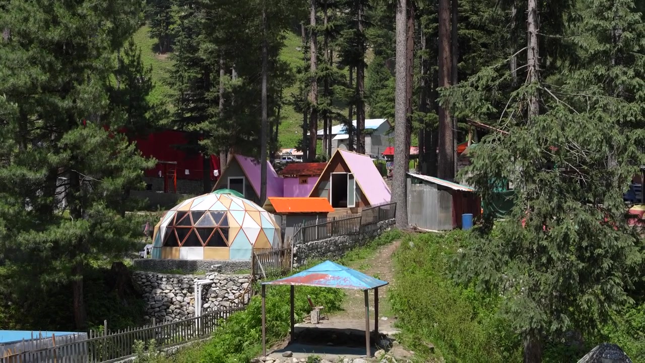 Colorful camping cabins and a geodesic dome stand among tall conifers in Kumrat Valley, Pakistan