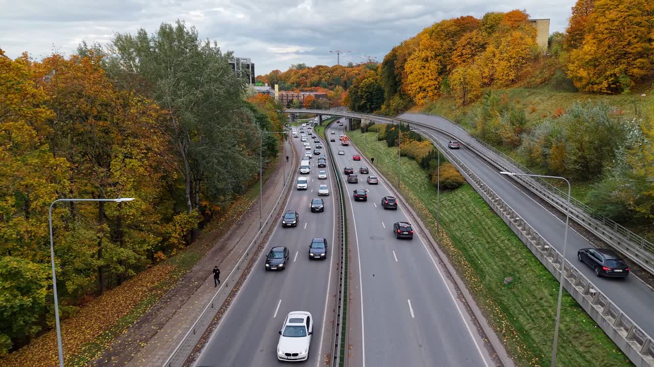 Dynamisk drone flyver fremad langs en flersporet vej i Vilnius, der følger den kørende trafik mellem efterårsskov og en fjern, moderne skyline.