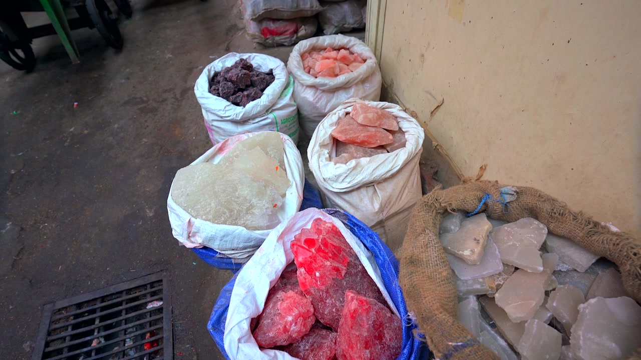 A high-angle static shot of traditional woven sacks filled with large raw blocks of pink Himalayan salt, white alum, and natural mineral crystals at a market stall in India