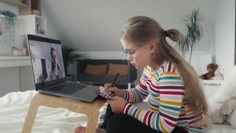 Kaukasisches-Mädchen-Im-Grundschulalter-Führt-Videogespräche-Auf-Dem-Laptop,-Während-Es-Auf-Dem-Bett-Sitzt.