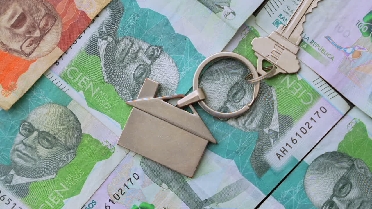 High-angle close-up of Colombian pesos and house-shaped keychain, symbolizing real estate investment. Bright lighting highlights crisp currency details. Ideal for finance themes