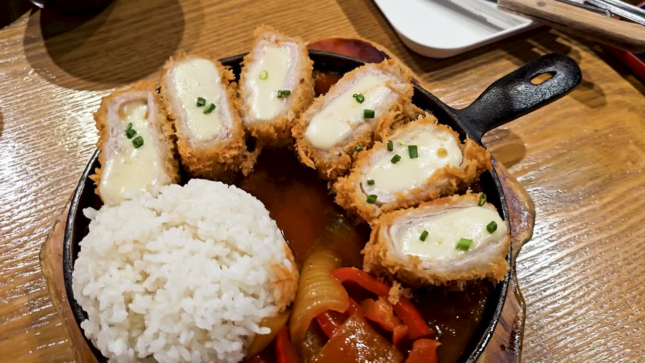 Golden crusted pork rolls stuffed with melted cheese and topped with green onion, presented alongside rice and veggies served in hot iron pan in Korean restaurant - parallax close-up