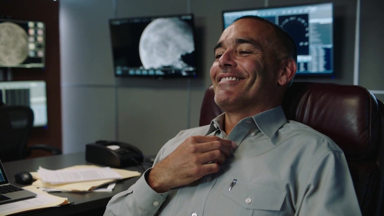 Smiling midlife man reacting to monitor alert, leaning back in leather chair, loosening gray shirt