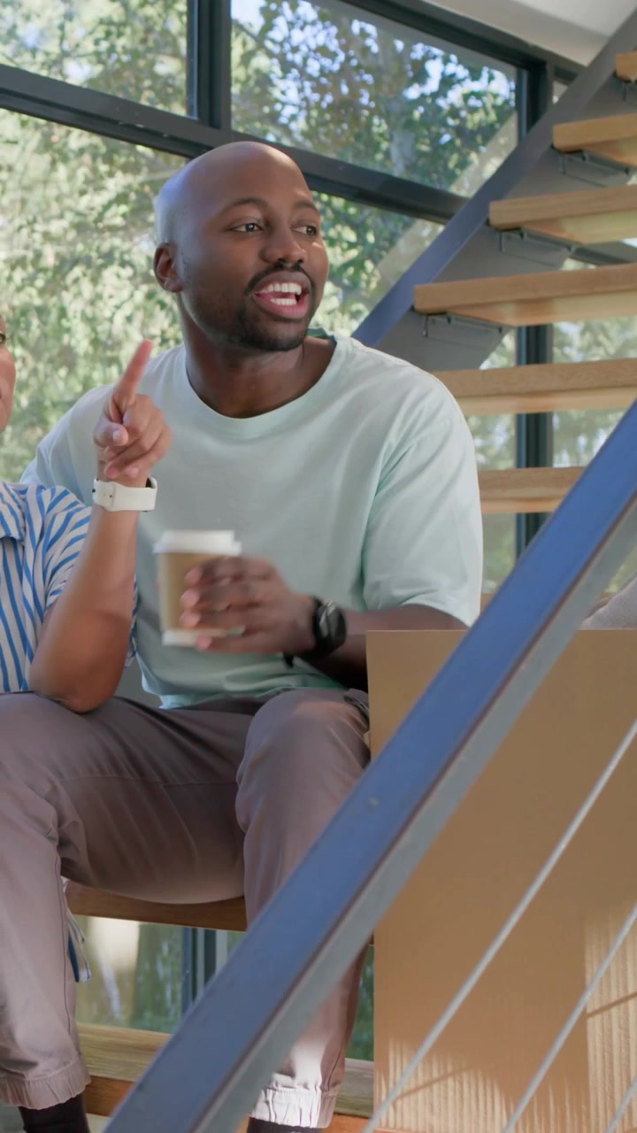 Vertical video: Laughing man reacting to chat reaching for moving box on stairs, holding coffee cup