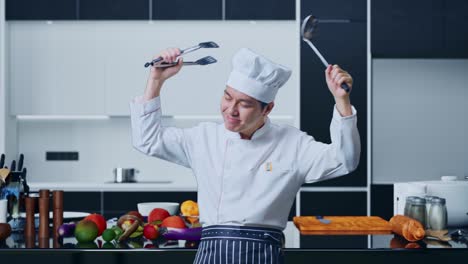 asian man chef holding tongs and ladle and dancing while standing in home kitchen