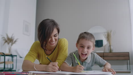 hermosa madre joven en un suéter cálido tendida en el suelo con mi hijo dibujando con marcadores en el papel retratando a su familia. el niño aprende a dibujar