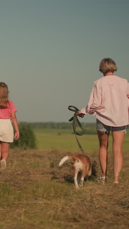 back view of pet lover walking with dog on leash and mother holding daughter s hand as they stroll through farmland, her hair blowing in wind, surrounded by lush greenery and distant trees
