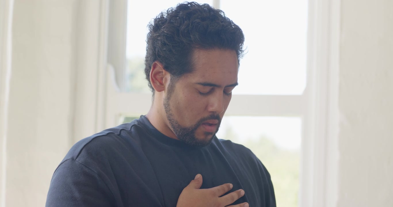 Uomo afroamericano in maglietta scura che si porta la mano destra al petto, respirando lentamente e calmandosi vicino alla finestra