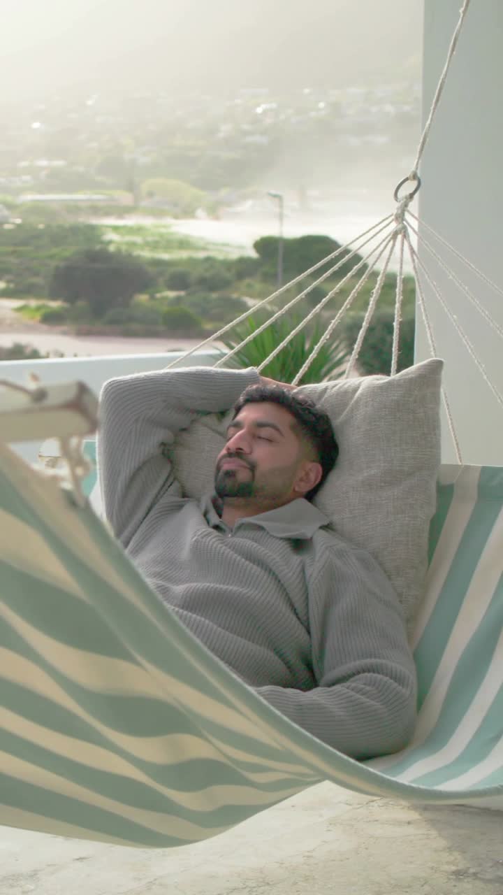 Video verticale: Un uomo sdraiato si muove sull'amaca sul balcone, raggiunge lo smartphone e legge i messaggi.