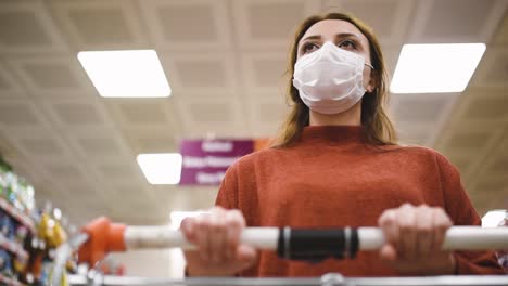 beautiful woman wearing protective disposable medical mask and fashionable clothes uses shopping cart while shopping in supermarket