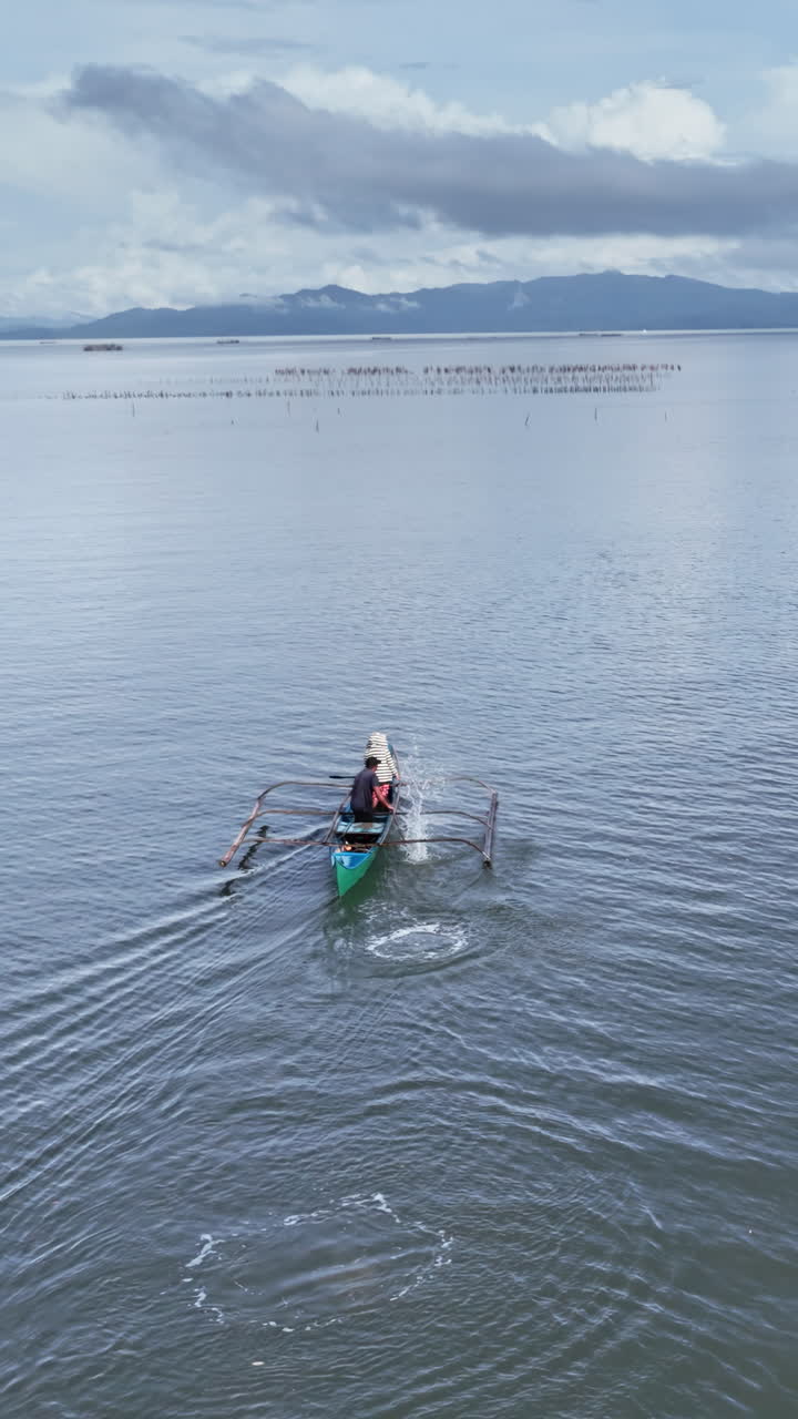 Aerial drone view of a fishermen rowing across calm open water with distant mountains and cloudy sky in the Philippines. Vertical